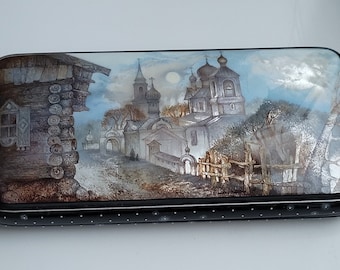 May include: A wooden box with a painted scene of a village with a church and a log cabin. The scene is painted in muted colors with a blue sky and white clouds. The box has a black lid with white polka dots.