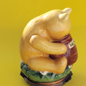 May include: A ceramic figurine of Winnie the Pooh, a honey bear, sitting on a green base with white flowers. He is holding a brown honey pot with the word "HUNNY" written on it.
