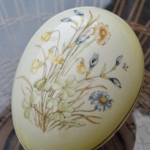 May include: A decorative, egg-shaped ceramic container with a floral design. The egg is cream-colored with a gold trim and features a bouquet of yellow, blue, and white flowers. The flowers are painted in a delicate, watercolor style.