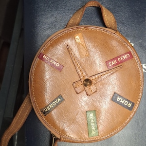 May include: A round, tan leather bag shaped like a clock face with two central hands. It features small rectangular labels in various colors printed with the Italian city names Milano, San Remo, Genova, Roma, and Firenze, along with a top handle.