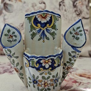 May include: A white ceramic vase with three compartments and a floral design. The vase has a blue and yellow trim, and the floral pattern includes red, blue, and green accents. The vase is likely handmade and suitable for displaying flowers.