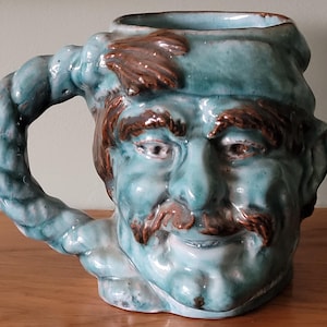May include: A ceramic mug shaped like a man's face with a rope handle. The mug is glazed in a light blue colour with brown accents for the hair, eyebrows, and moustache.