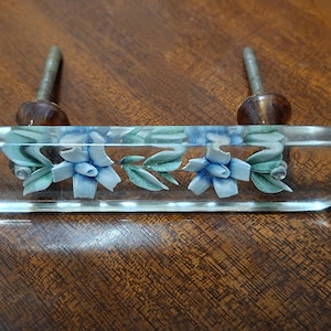 May include: A clear glass cabinet pull with a rectangular shape. The pull features a floral design with blue and green painted flowers and leaves. The hardware has two metal screws with brown accents.