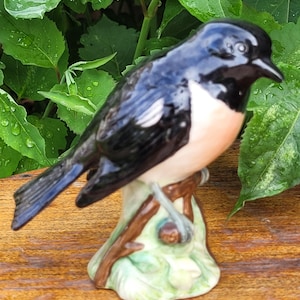 May include: A ceramic figurine of a black and pink bird perched on a branch. The bird has a white patch on its wing and is sitting on a green and brown base.