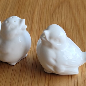 May include: Two white ceramic bird salt and pepper shakers. The birds are facing each other, with one bird's head tilted up and the other bird's head tilted down.
