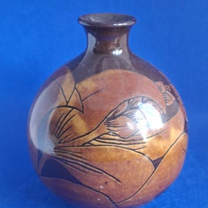 May include: A small, round, brown ceramic vase with a narrow neck and a glossy finish. The surface features dark, etched line drawings of large, stylized floral petals and leaves that wrap around the spherical body of the vessel.