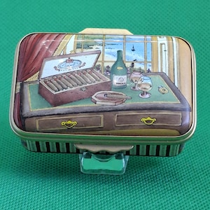 May include: A rectangular, hinged metal box with a painted scene of a desk with cigars, a bottle of wine, and glasses. The box has a gold-colored trim and a green interior. The background shows a window with a view of the sea.