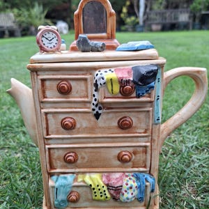 May include: A whimsical teapot shaped like a dresser, with five drawers and a handle. The teapot is beige with brown drawer knobs and colorful clothing draped over the drawers. A small clock and mirror sit on top.
