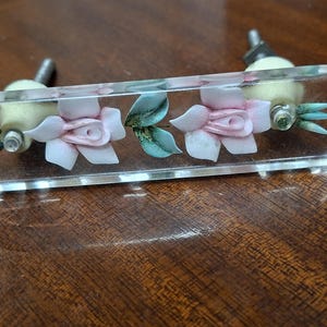 May include: A clear rectangular cabinet pull with a floral design. The pull features two pink flowers with green leaves and stems encased in clear acrylic. The hardware is silver-toned.