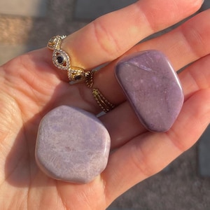 May include: Two smooth, polished purple stones rest in an open hand, alongside a gold ring with black and clear stone accents. The stones have a soft, matte finish and vary slightly in shape. The ring is intricately designed.