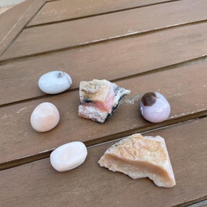May include: Assortment of polished gemstones and raw mineral specimens in shades of pink, white, and tan. The collection includes oval and irregular shapes, displayed on a wooden surface. Ideal for crystal collectors and those interested in natural stones.