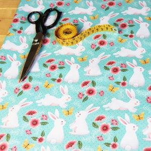 Peut inclure: Tissu turquoise avec un motif répété de lapins blancs, de fleurs roses et de papillons jaunes. Un mètre ruban jaune et des ciseaux à tissu noirs sont posés sur le tissu.