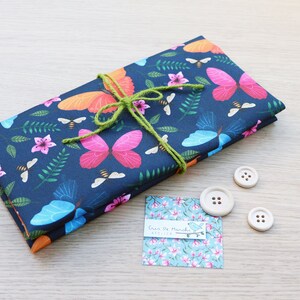 May include: A stack of fabric with a butterfly and floral print in shades of blue, pink, orange, and green. The fabric is tied with a green ribbon and has a small tag that says "Cric'o de Manchu Atelier".