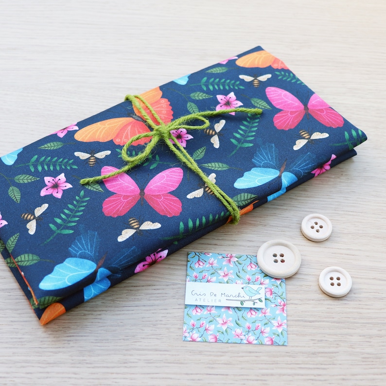May include: A stack of fabric with a butterfly and floral print in shades of blue, pink, orange, and green. The fabric is tied with a green ribbon and has a small tag that says "Cric'o de Manchu Atelier".