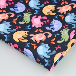 Op de afbeelding: Donker marineblauwe stof met een herhaald patroon van kleurrijke cartoonkatten en vlinders. De katten hebben verschillende poses en kleuren, waaronder roze, blauw, geel en groen. De vlinders zijn oranje en roze.