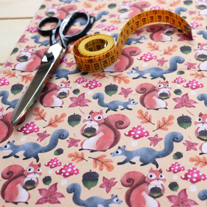May include: A beige fabric with a repeating pattern of squirrels holding acorns. The squirrels are red, brown, and gray. There are also leaves, acorns, and mushrooms in the pattern. A yellow measuring tape and a pair of silver scissors are on top of the fabric.