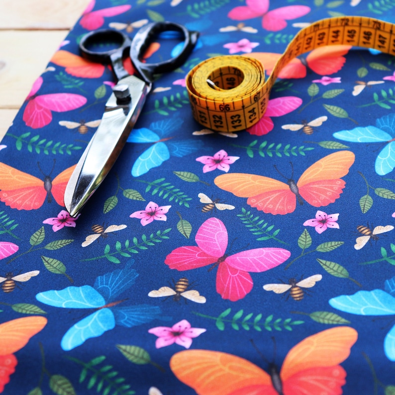 May include: A blue fabric with a repeating pattern of butterflies, flowers, and bees. A yellow measuring tape and a pair of silver scissors are lying on top of the fabric.