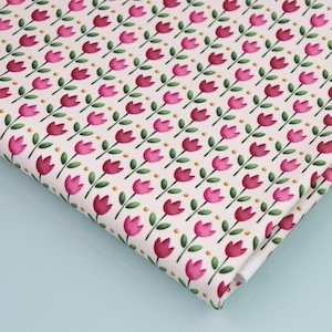 Può includere: Tessuto piegato con un motivo ripetuto di fiori di tulipano rosa, steli e foglie verdi e piccoli punti dorati su sfondo crema. Il motivo floreale è disposto diagonalmente sul tessuto.