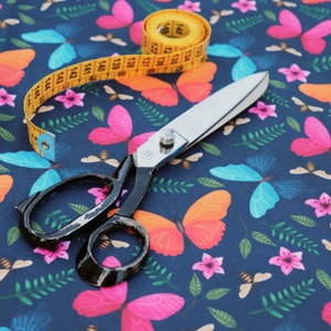 May include: A black fabric scissors with a yellow measuring tape on a blue background with a butterfly pattern. The measuring tape has numbers printed on it, including 133, 134, 135, 136, 137, 138, 139, 140, 141, 142, 143, 144, 145, and 146.