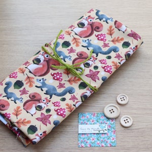 May include: A beige fabric with a repeating pattern of squirrels, acorns, and fall leaves. The fabric is tied with a green ribbon and there are three wooden buttons on a wooden surface.