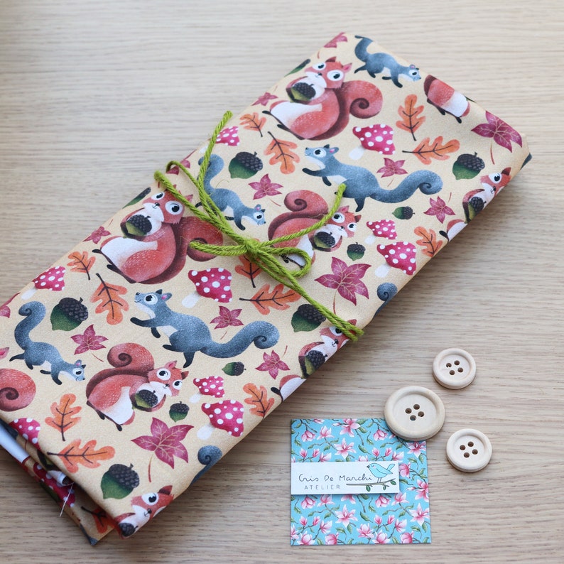 May include: A beige fabric with a repeating pattern of squirrels, acorns, and fall leaves. The fabric is tied with a green ribbon and there are three wooden buttons on a wooden surface.