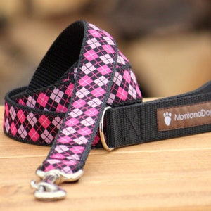 May include: A pink and black plaid dog leash with a silver clip and a brown leather patch with the text "MontanaDog" on it.