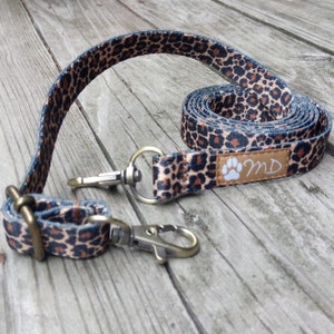Puede incluir: Una correa para perros enrollada con un estampado de leopardo en marrón y negro. La correa tiene un cierre metálico y un pequeño parche con las iniciales "MD" y una huella de pata. Está sobre una superficie de madera.