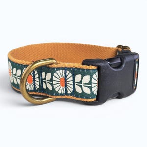 May include: A pet collar featuring a dark green fabric strap with a repeating white and orange floral pattern. The collar includes a mustard yellow inner lining, a brass-toned metal D-ring, and a black plastic side-release buckle closure.