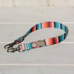 May include: A colorful dog leash with a striped pattern in teal, orange, and red. The leash has two metal clasps and a small tag with a paw print and the initials "MD". The background is a light wood.