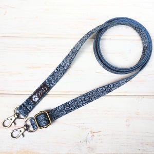 Puede incluir: Una correa para perros con estampado azul y blanco con cierres metálicos y una hebilla dorada. La correa tiene un diseño geométrico repetitivo y las iniciales "MD" con una huella de pata.