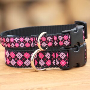 May include: Two dog collars with a black background and a pink and black argyle pattern. Each collar has a black plastic buckle and a silver metal D-ring. The collars are made of a durable fabric.