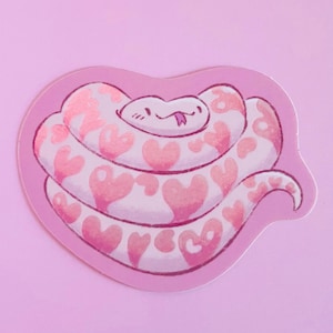 Op de afbeelding: Een roze sticker met een cartoon slang opgerold in een hartvorm. De slang is wit met roze hartpatronen. De sticker heeft een roze rand en staat op een roze achtergrond.