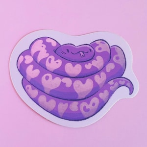 Op de afbeelding: Een sticker met een cartoon slang opgerold in een hartvorm. De slang is paars met lichtroze hartvormige markeringen. De sticker heeft een witte rand en staat tegen een lichtroze achtergrond.