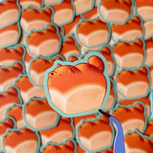 Op de afbeelding: Een verzameling stickers met een cartoon Shiba Inu-hond die is ontworpen om op een brood te lijken. De stickers hebben een lichtblauwe rand en zijn oranje en wit. Een blauwe pincet houdt een sticker vast.