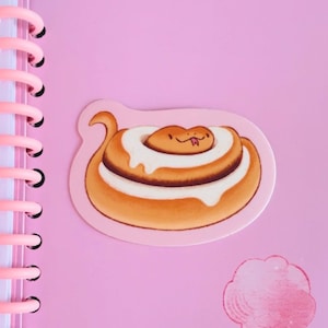 May include: A sticker featuring a cartoon snake in the shape of a cinnamon roll with white icing. The sticker is on a pink background with a spiral notebook visible on the left side.