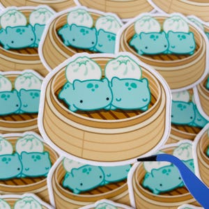 May include: A set of stickers featuring cartoon dumplings in a bamboo steamer. The dumplings are light blue with white accents and are arranged in a traditional dim sum style.