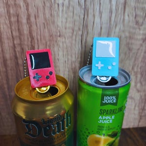 以下が含まれることがあります： 赤と青の2つのミニチュアゲームコンソールのキーホルダーが、開いた飲料缶の上に置かれています。赤いコンソールには黒い画面と十字型のボタンがあります。青いコンソールにはプラス記号とドットがあります。