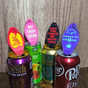 May include: A collection of novelty keychains in various colors, including red, pink, black, and purple, each with a unique phrase. The keychains are placed on top of beverage cans, such as sparkling apple juice and Dr. Pepper.