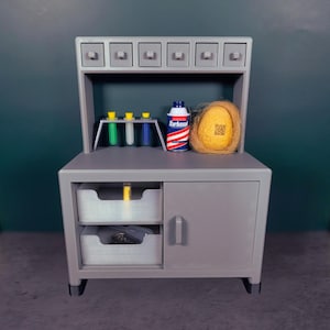 May include: A grey miniature laboratory set with a cabinet, drawers, and a shelf. The shelf holds test tubes, a shaving cream can, and a yellow object. The cabinet has two open trays and a closed door.