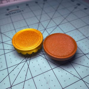 May include: Two circular objects with textured surfaces. One is gold with a scalloped edge, the other is reddish-brown. They are on a blue cutting mat. The objects have a raised central area. The gold object has a yellow scalloped edge.