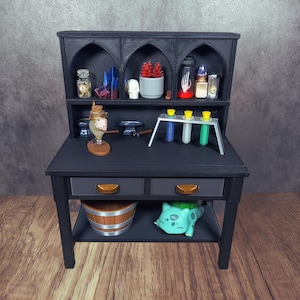 Può includere: Una scrivania nera in miniatura con una credenza, con vari piccoli oggetti. La credenza ha ripiani ad arco con bottiglie, cristalli e un teschio. La scrivania ha cassetti, una botte e una figura verde di Bulbasaur.