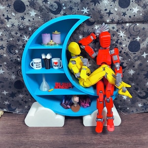 Pode incluir: Uma prateleira azul em forma de meia-lua com três níveis, contendo itens em miniatura e duas figuras de robôs. A prateleira contém pequenos potes, canecas com texto e outras peças decorativas. Os robôs são vermelhos e amarelos. A prateleira fica em uma base de nuvem branca.