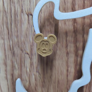 May include: A light brown Mickey Mouse-shaped charm with a smiling face and two large ears. The charm is attached to a white, curved decorative element. The background is a wooden surface.