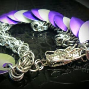 May include: A silver chainmail bracelet with a dragon head charm and purple and white scales. The scales are made of metal and have a gradient effect.