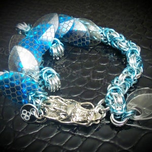 May include: A silver chain bracelet with blue and silver scales and a silver dragon charm. The bracelet is on a black background.
