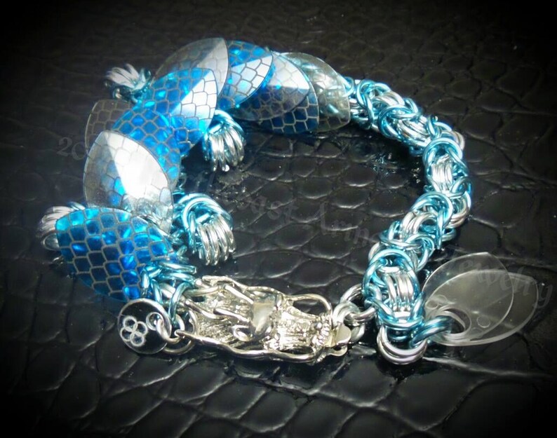 May include: A silver chain bracelet with blue and silver scales and a silver dragon charm. The bracelet is on a black background.