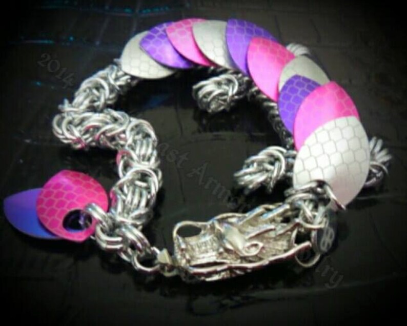 May include: A silver chainmail bracelet with a dragon head charm. The bracelet is decorated with pink, purple, and white scales.