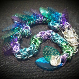 May include: A multi-colored chainmail bracelet with iridescent purple, blue, and green scales. The bracelet has a silver clasp and a small silver charm.