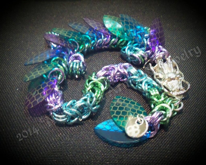 May include: A multi-colored chainmail bracelet with iridescent purple, blue, and green scales. The bracelet has a silver clasp and a small silver charm.
