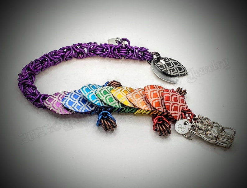 May include: A rainbow dragon scale bracelet with a purple chain and a silver dragon head charm. The scales are made of metal and have a white and gray pattern.